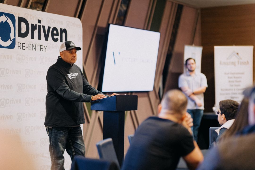 A man is giving a presentation in front of a sign that says driven