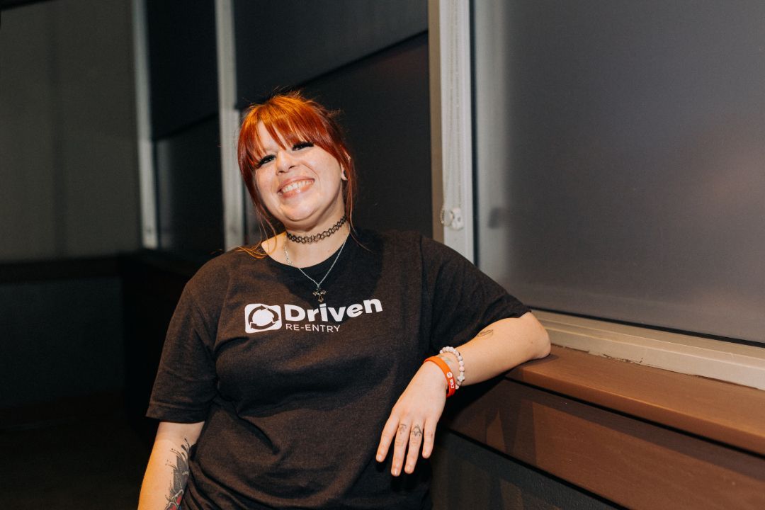 A woman wearing a black driven t-shirt is leaning against a wall and smiling.