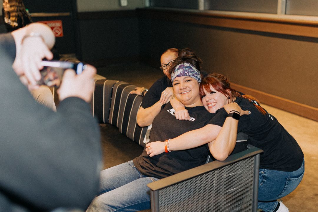 A man is taking a picture of two women hugging each other on a couch.