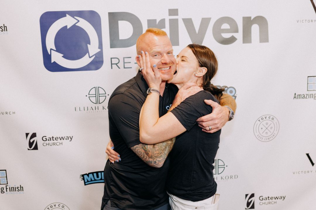 A man and a woman are hugging and kissing in front of a sign that says driven.