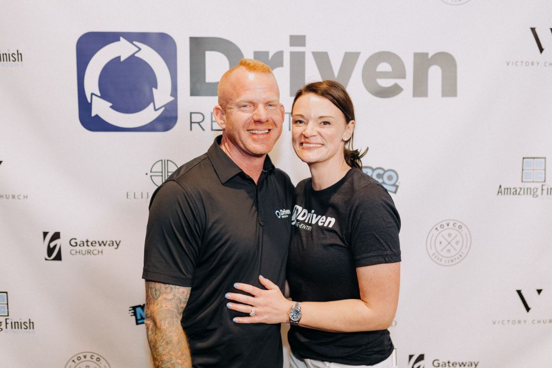 A man and a woman are posing for a picture in front of a sign that says driven.