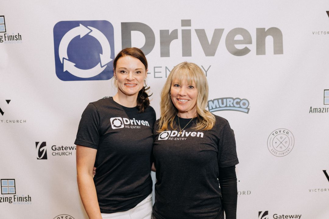 Two women are posing for a picture in front of a sign that says driven.
