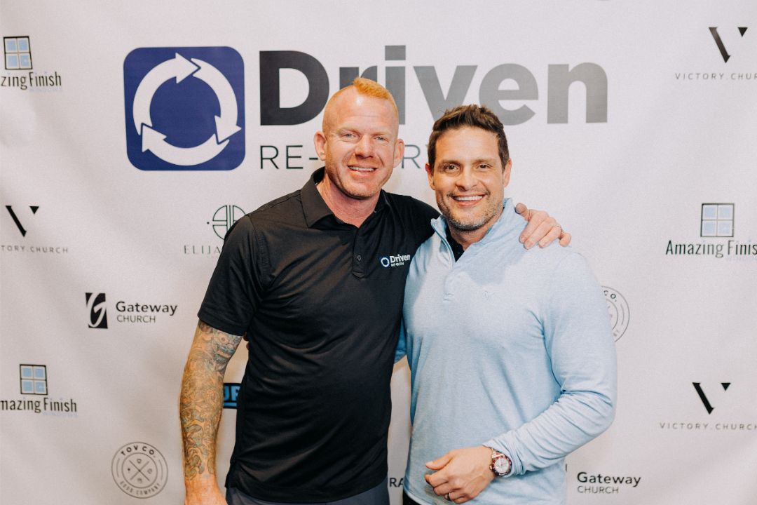 Two men are posing for a picture in front of a sign that says driven.