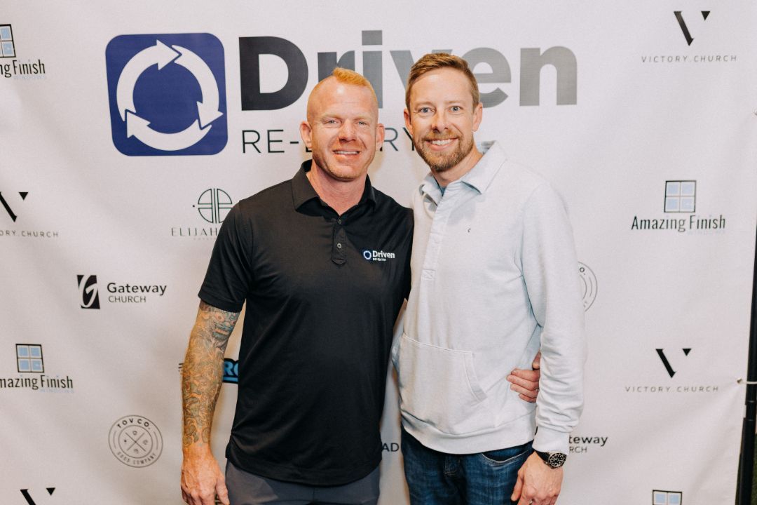 Two men are posing for a picture in front of a sign that says driven.