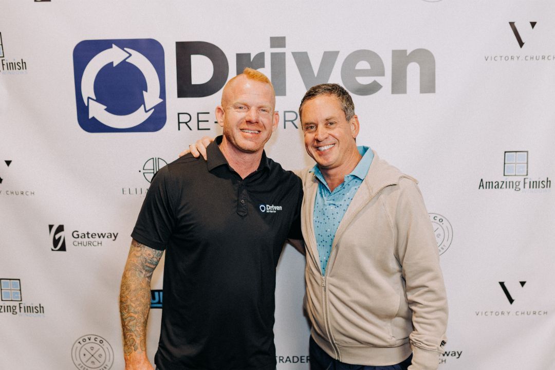 Two men are posing for a picture in front of a sign that says driven.