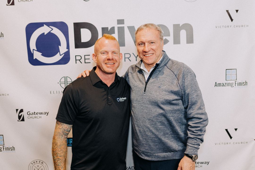 Two men are posing for a picture in front of a sign that says driven recovery.