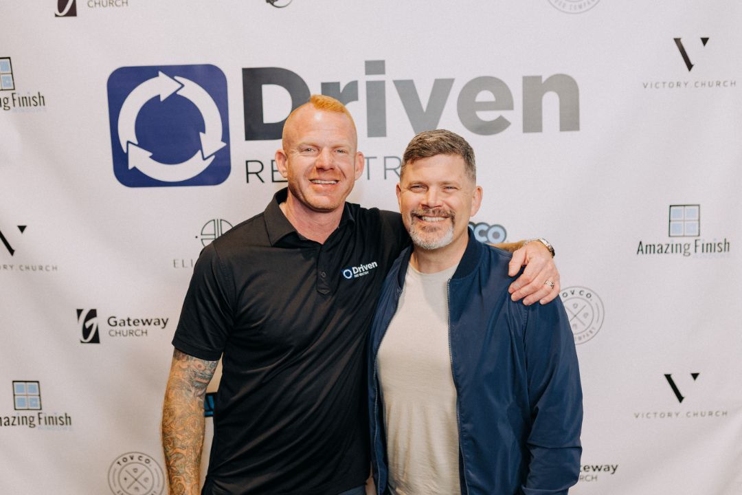 Two men are posing for a picture in front of a sign that says driven.