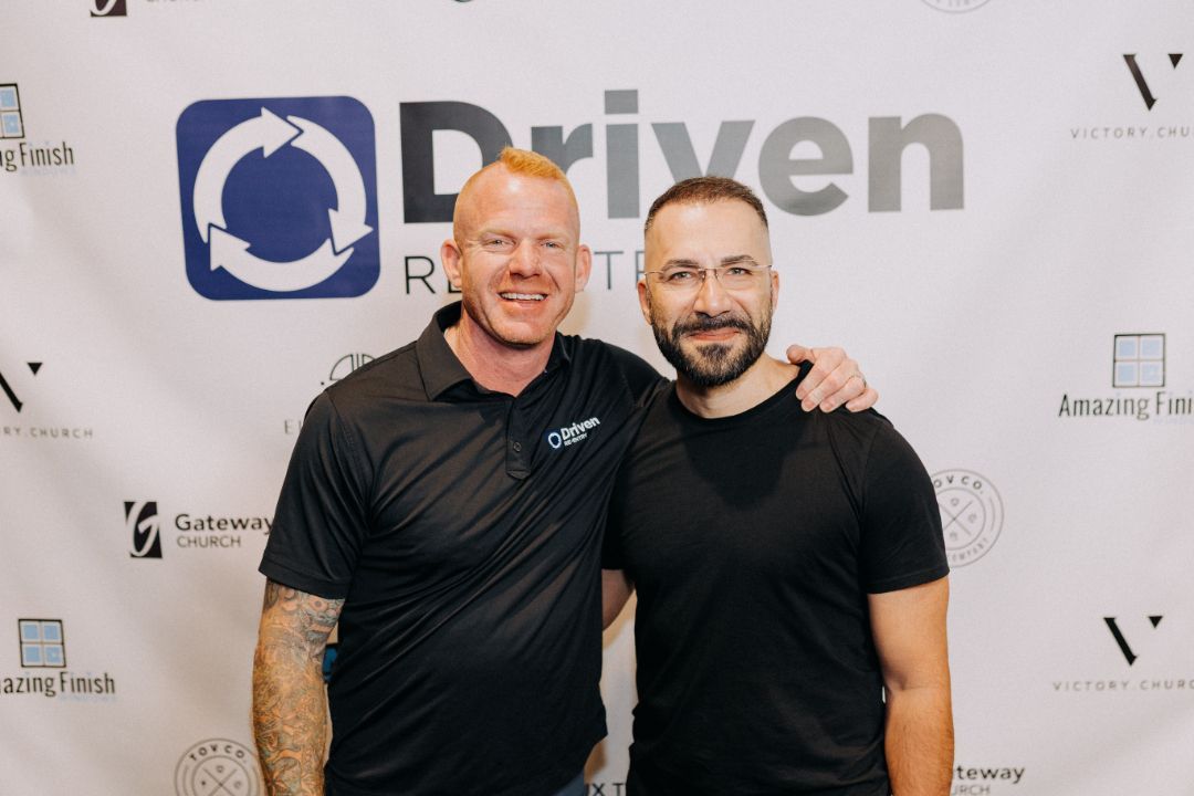 Two men are posing for a picture in front of a sign that says driven.