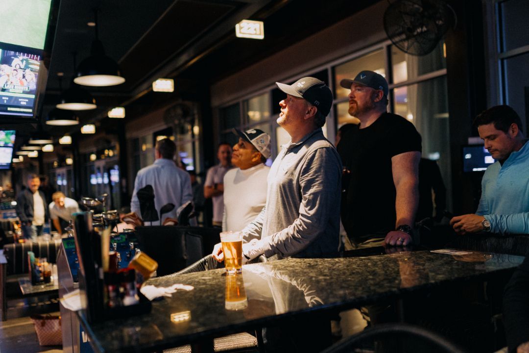 A group of men are standing at a bar watching a game.
