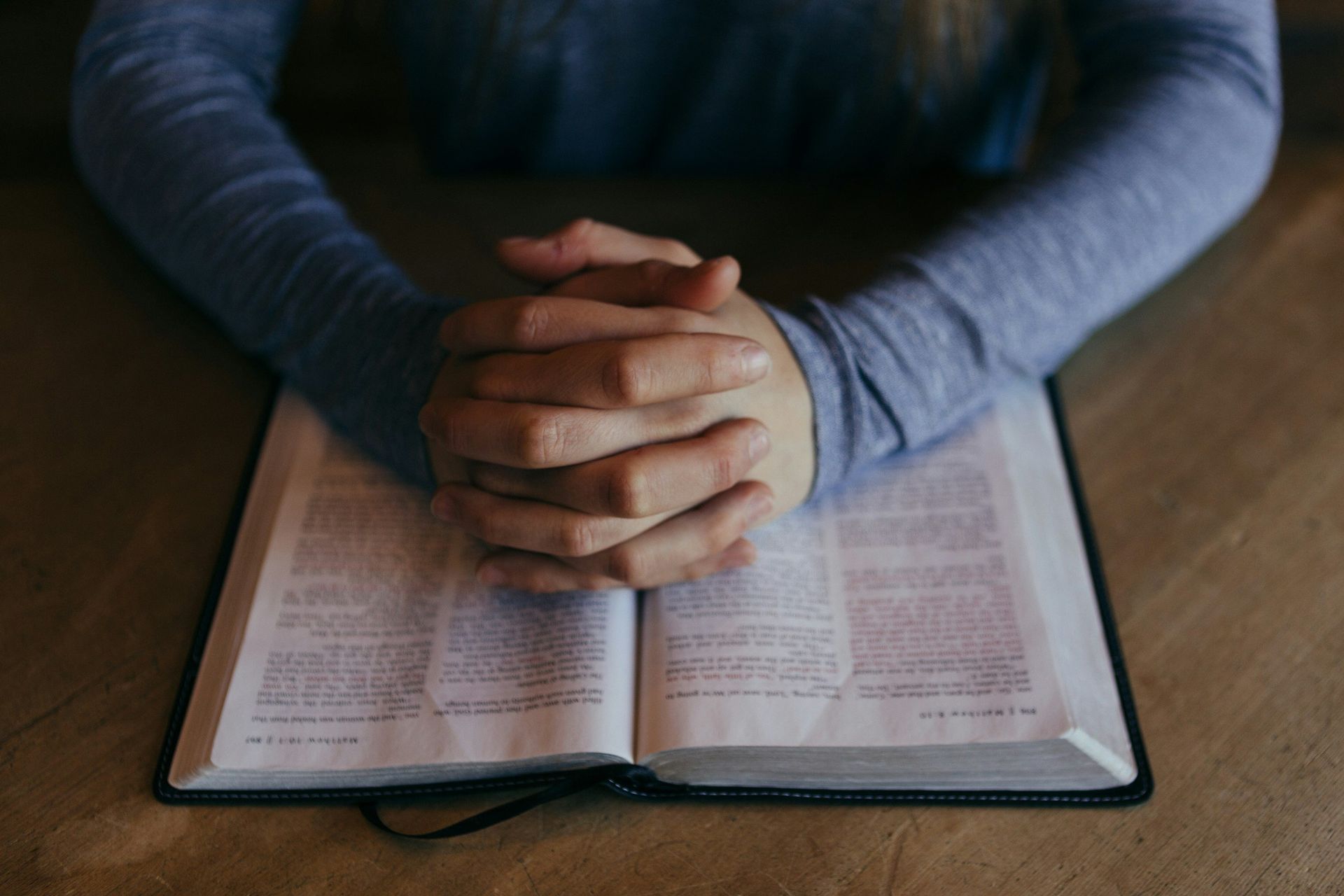 A man is sitting at a table with her hands folded over an open bible.
