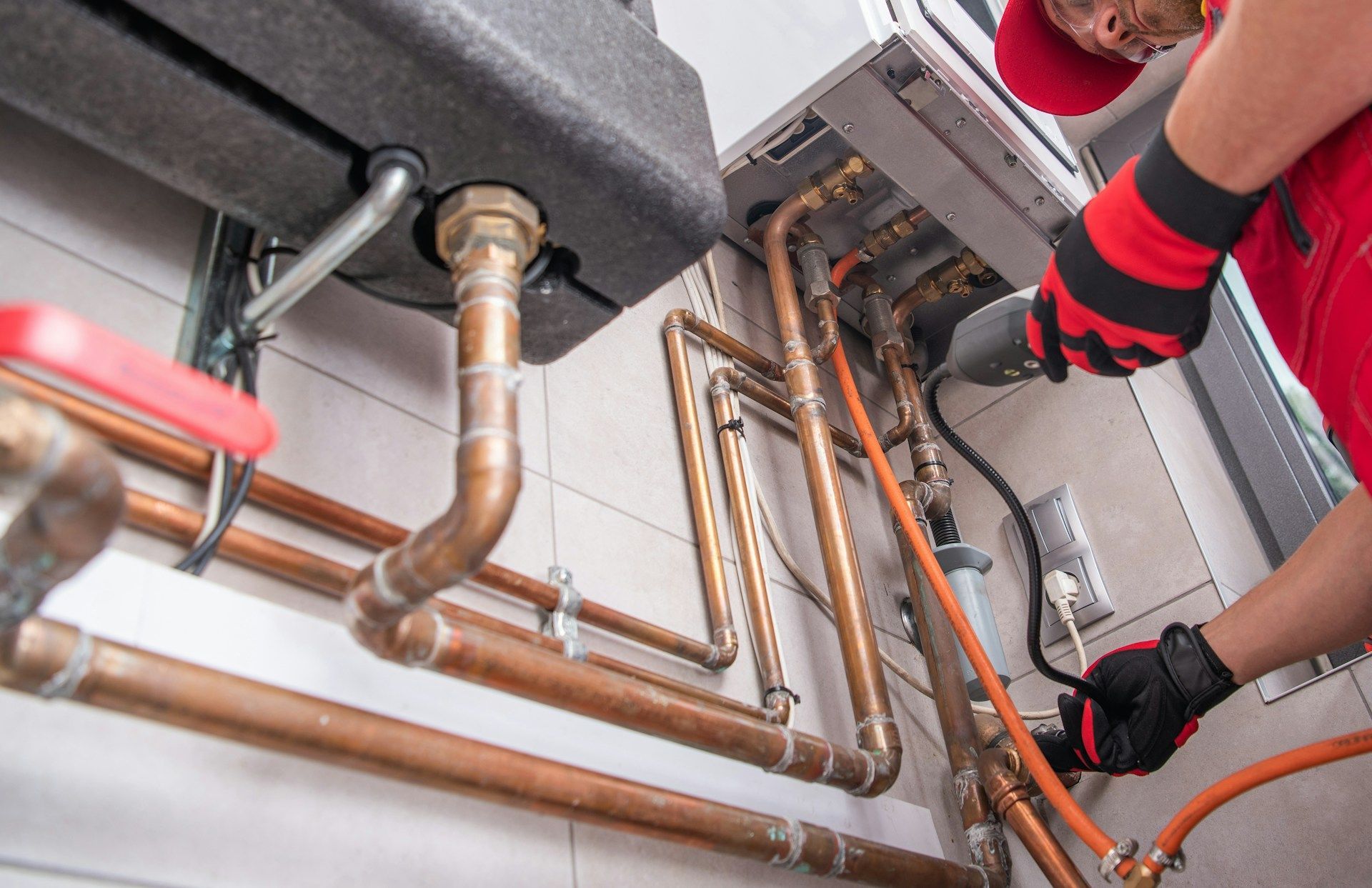 Technician inspecting exposed copper pipes and heating system in a utility room, wearing red gloves.
