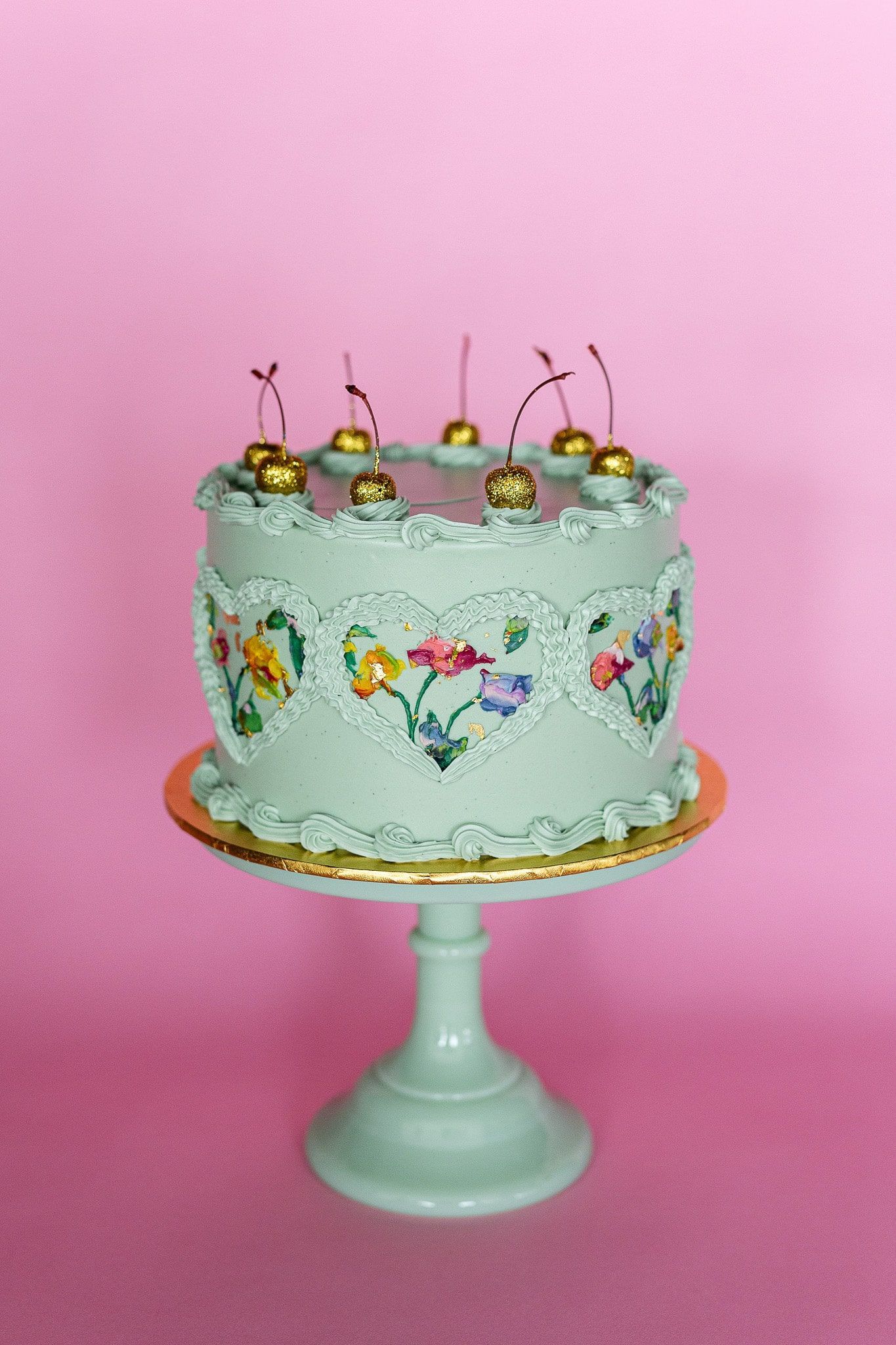 A floral-decorated cake with cherries on a pedestal, set against a pink backdrop.