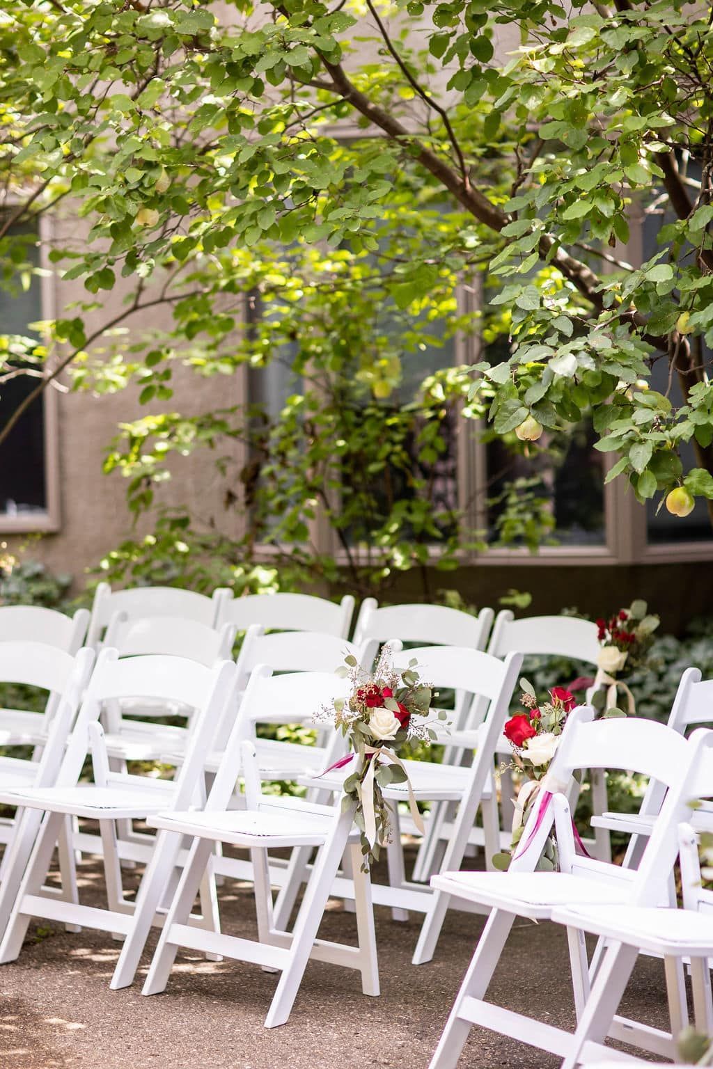 A row of white folding chairs with flowers on them are sitting under a tree.