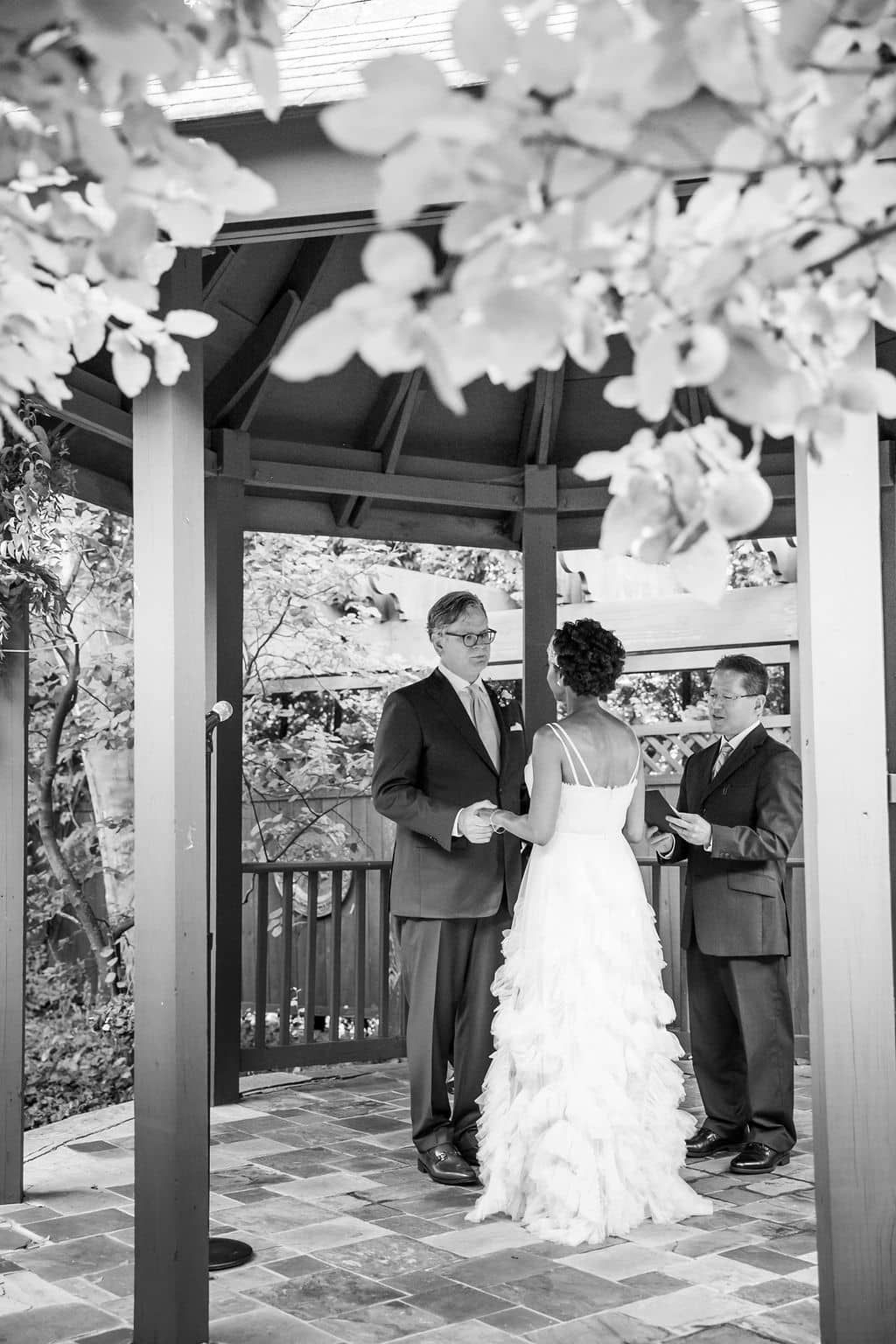 A black and white photo of a bride and groom holding hands under a gazebo.