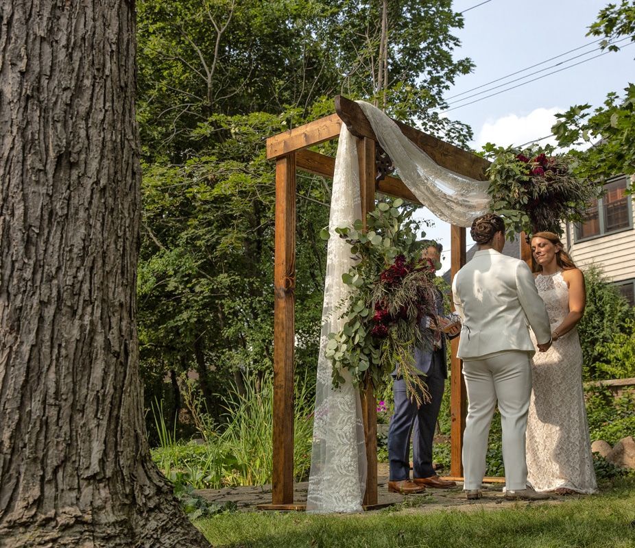 A bride and groom are standing under a wooden arch decorated with flowers.