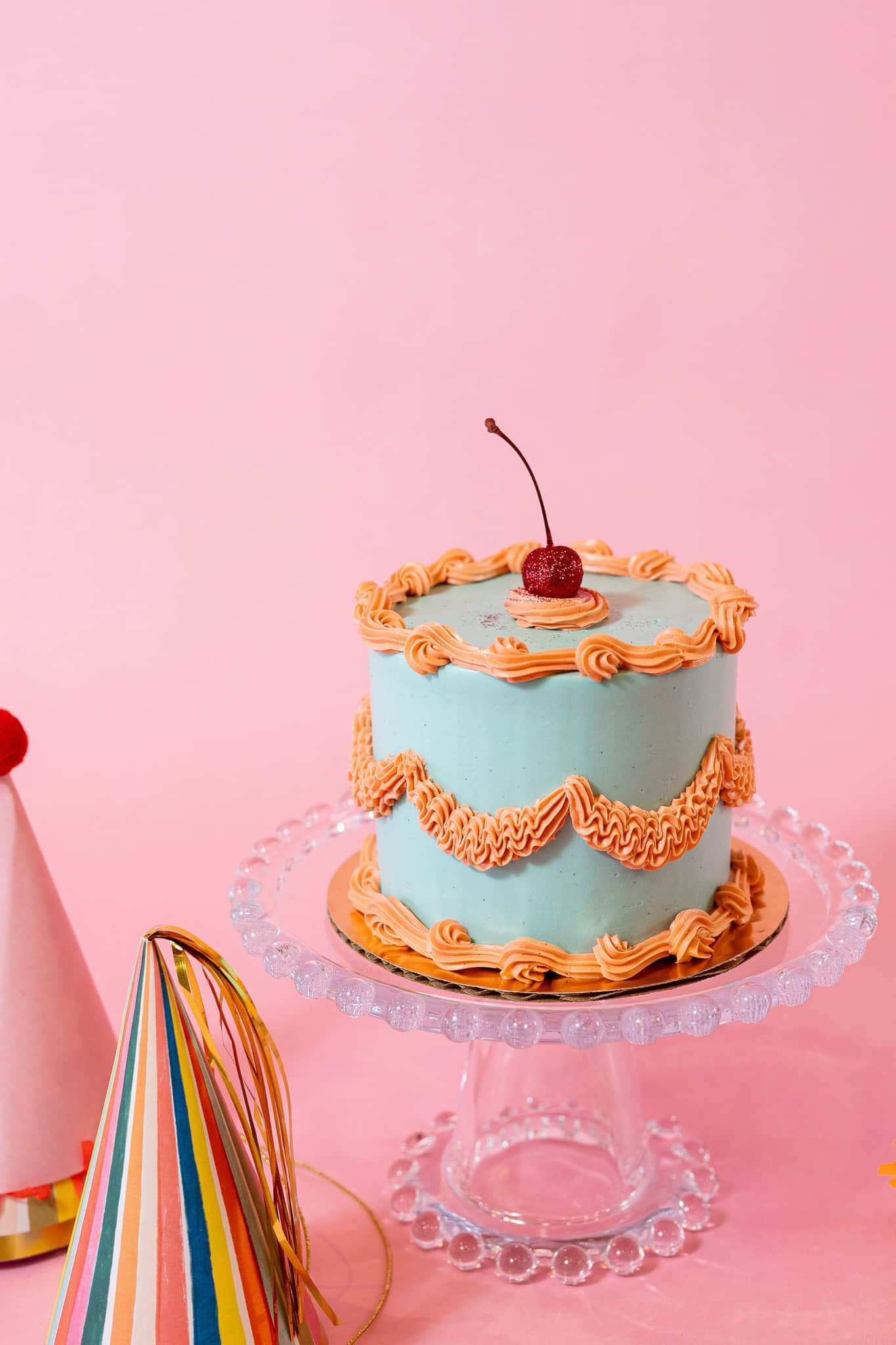 Blue frosted cake with orange trim, topped with a cherry, on a glass cake stand, next to party hats on pink background.