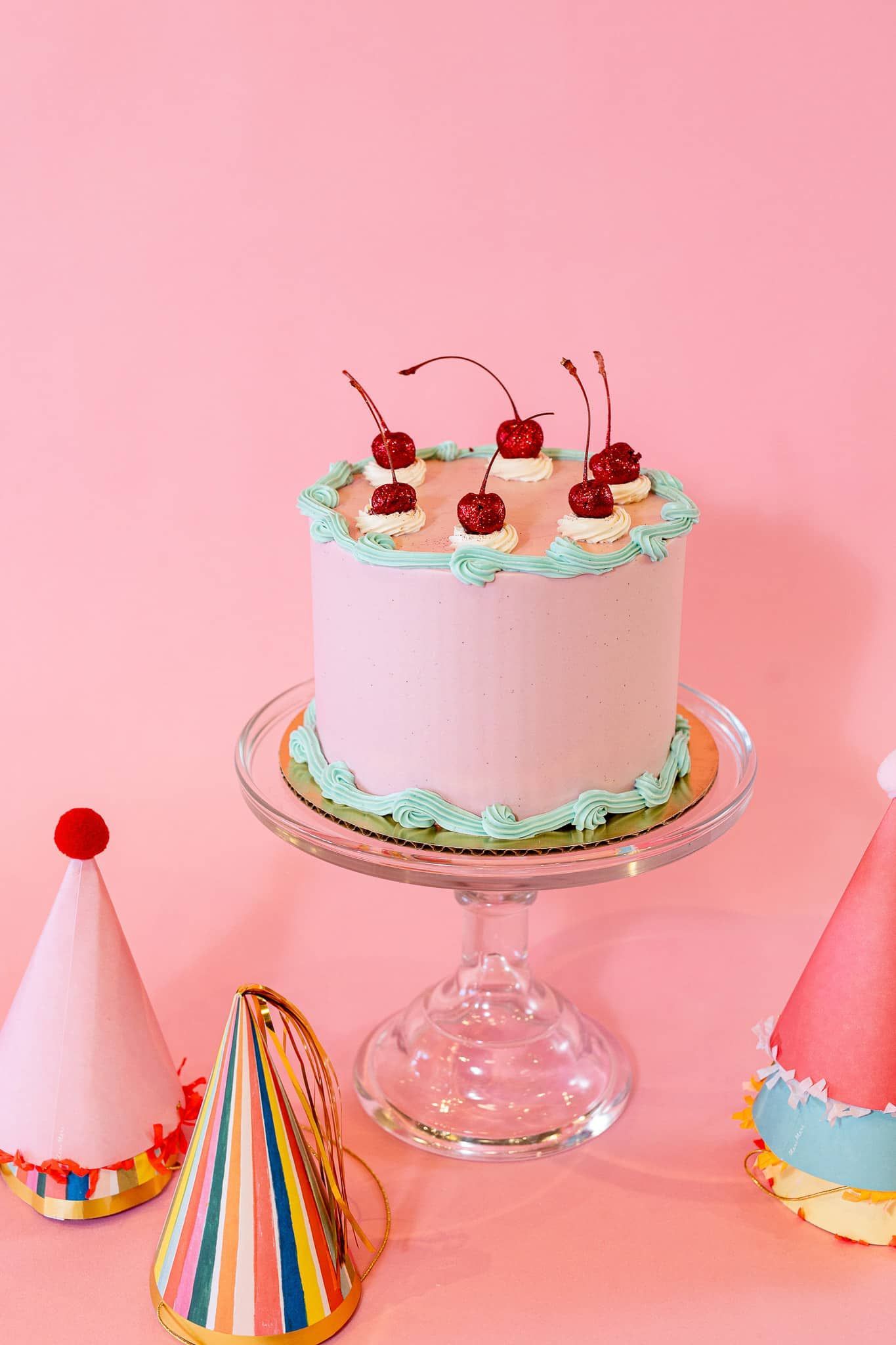 Pink cake with cherries on glass stand, surrounded by party hats on pink background.
