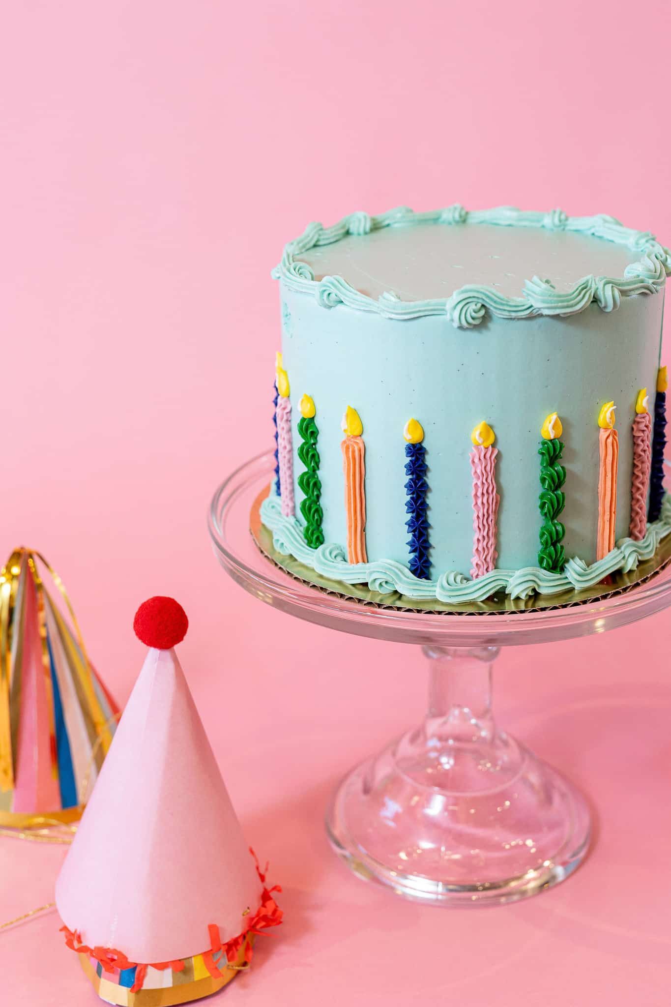 Birthday cake with colorful candles on a glass cake stand, party hats on pink backdrop.