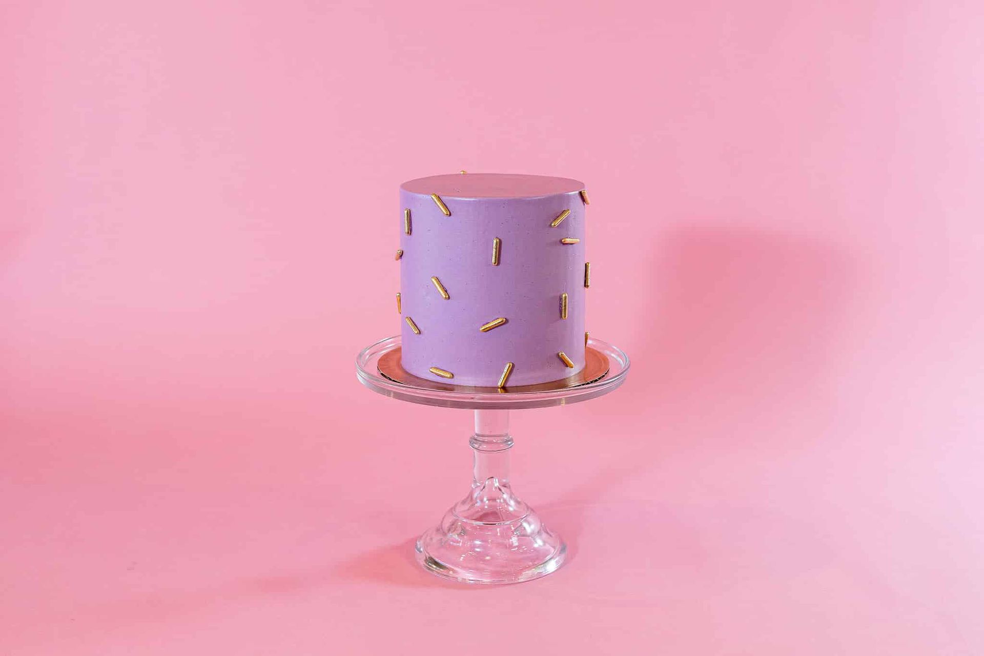 Purple cake on a glass cake stand against a pink background, decorated with gold sprinkles.