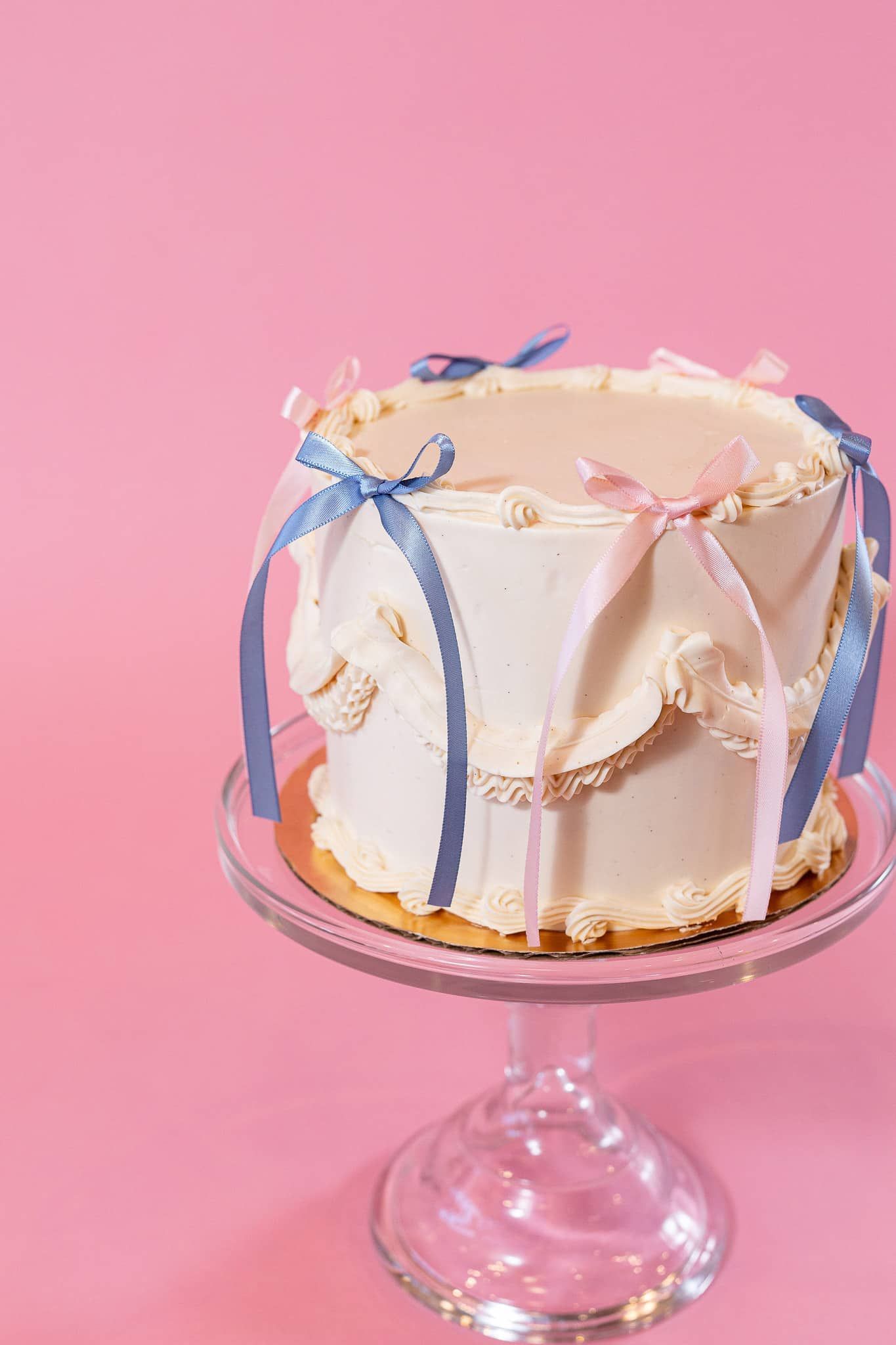 Cake on glass stand decorated with blue and pink ribbons against pink backdrop.