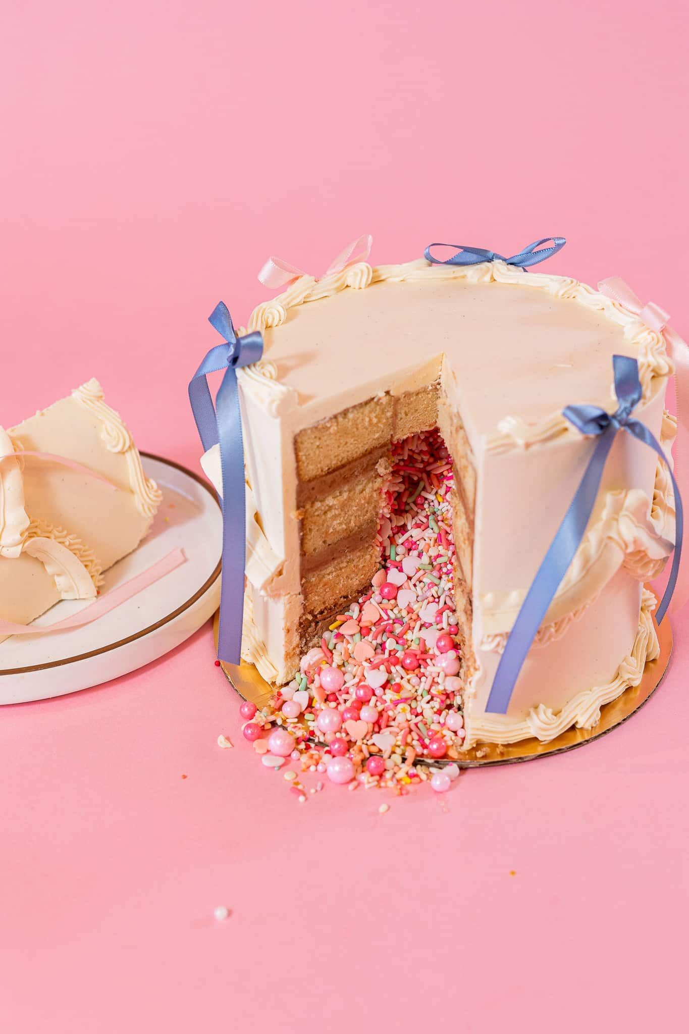 Cake with frosting, a slice removed revealing colorful sprinkles pouring out. Pink background.
