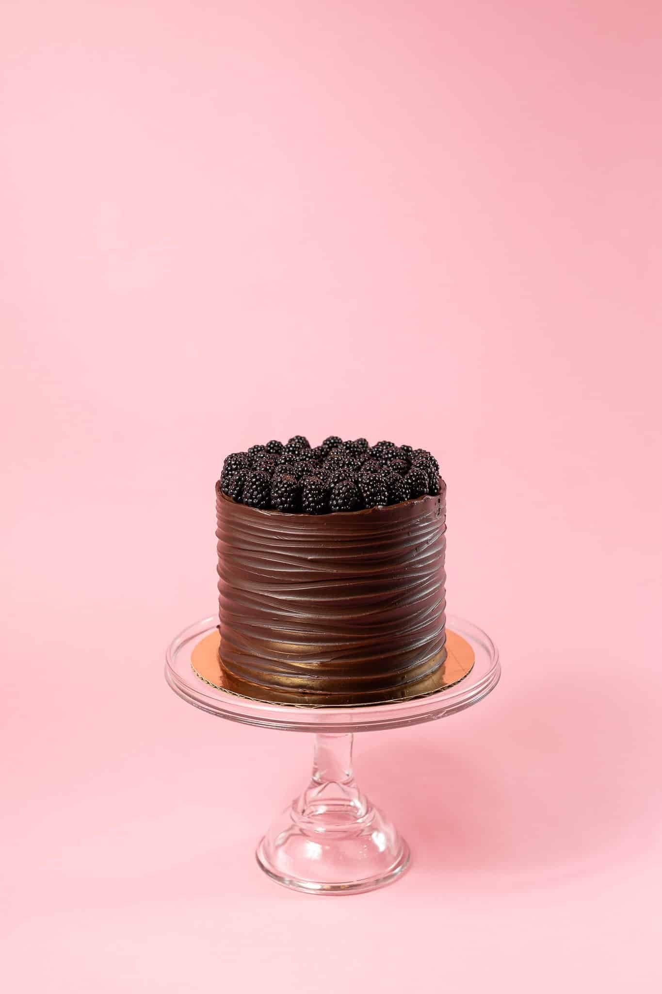 Chocolate cake with dark frosting and berries on glass cake stand, pink backdrop.