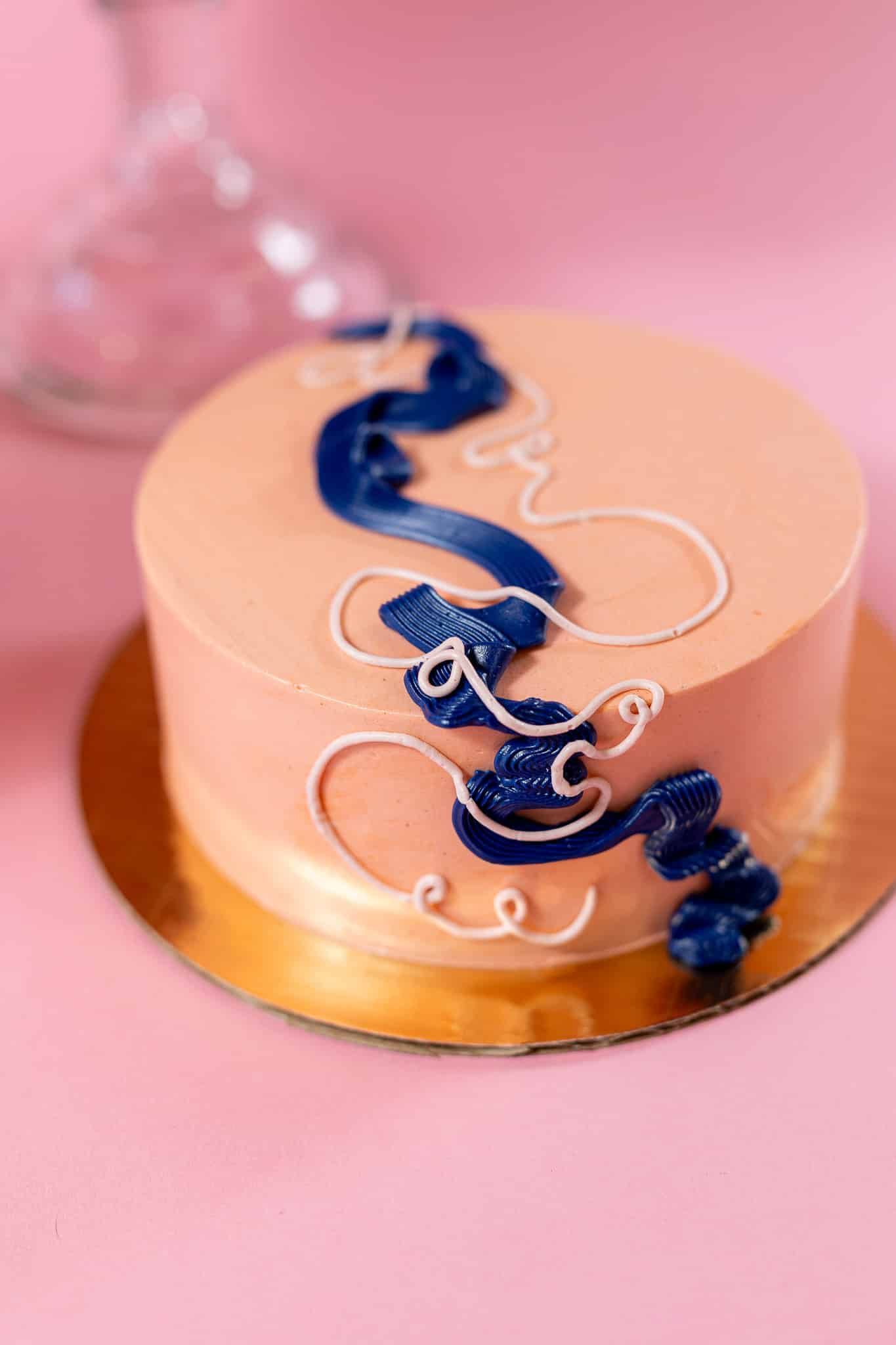 Round peach cake with blue and white frosting decorations on a gold cake board on a pink surface.