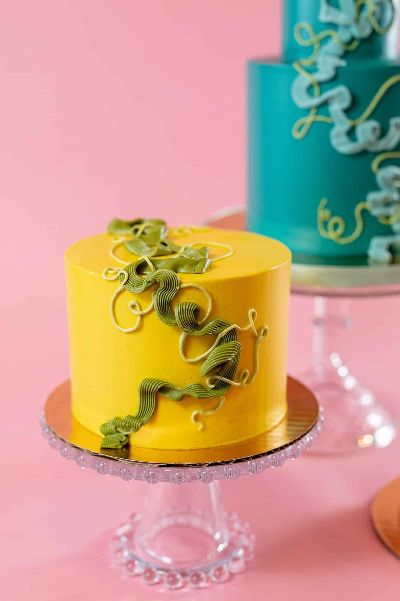 Yellow cake on a clear stand, decorated with green swirls. Pink background.