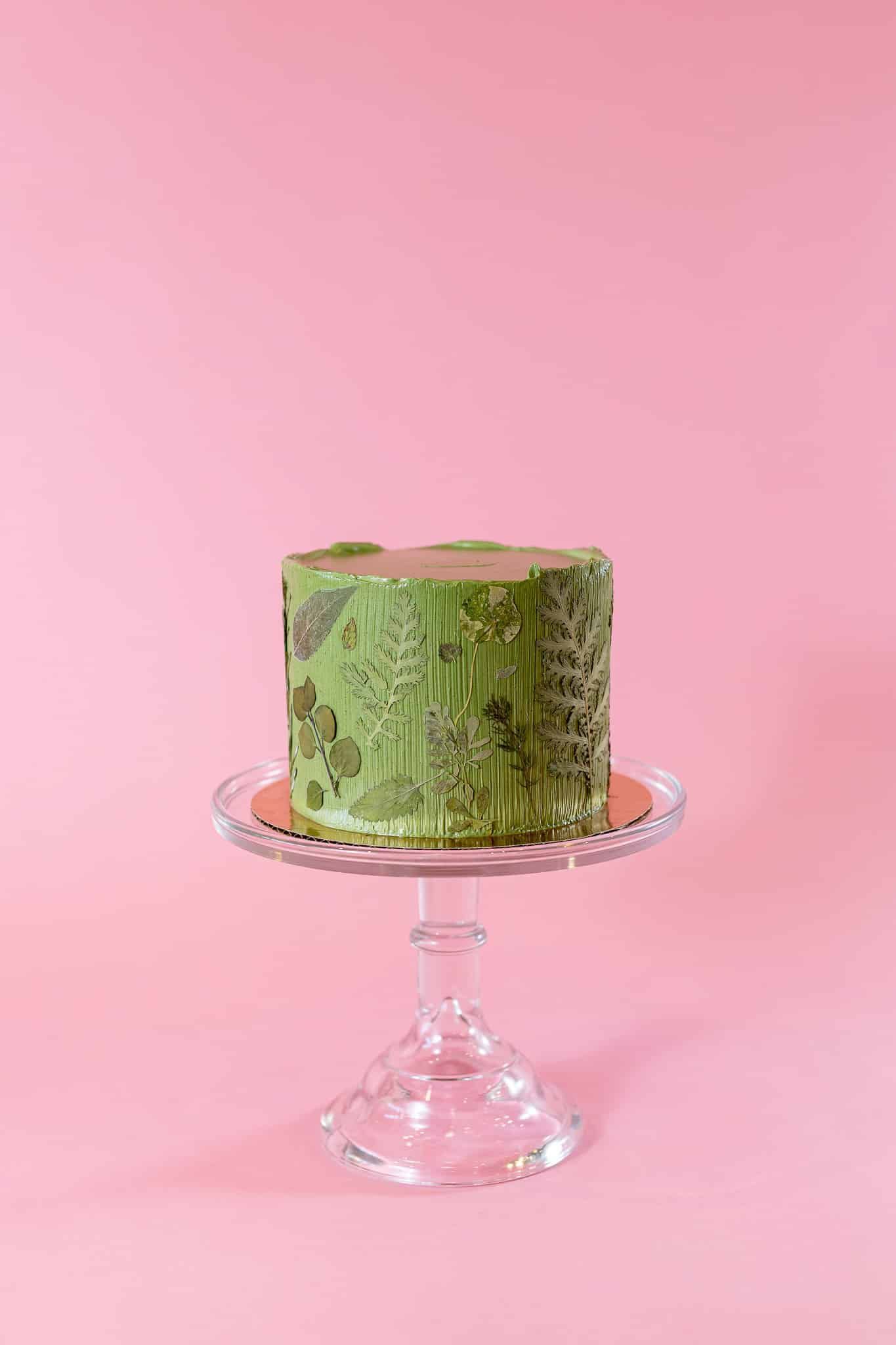 Green frosted cake on a glass cake stand, set against a pink background.