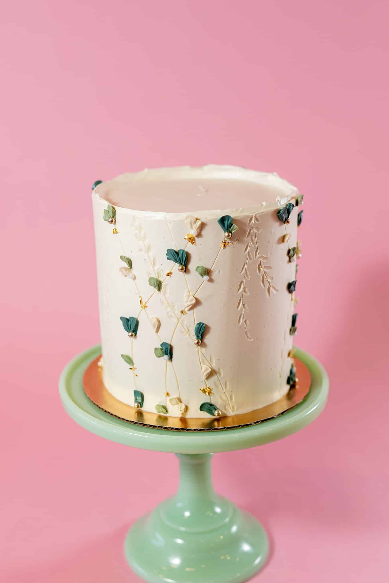 White frosted cake on a green stand, decorated with blue and gold accents, against a pink backdrop.