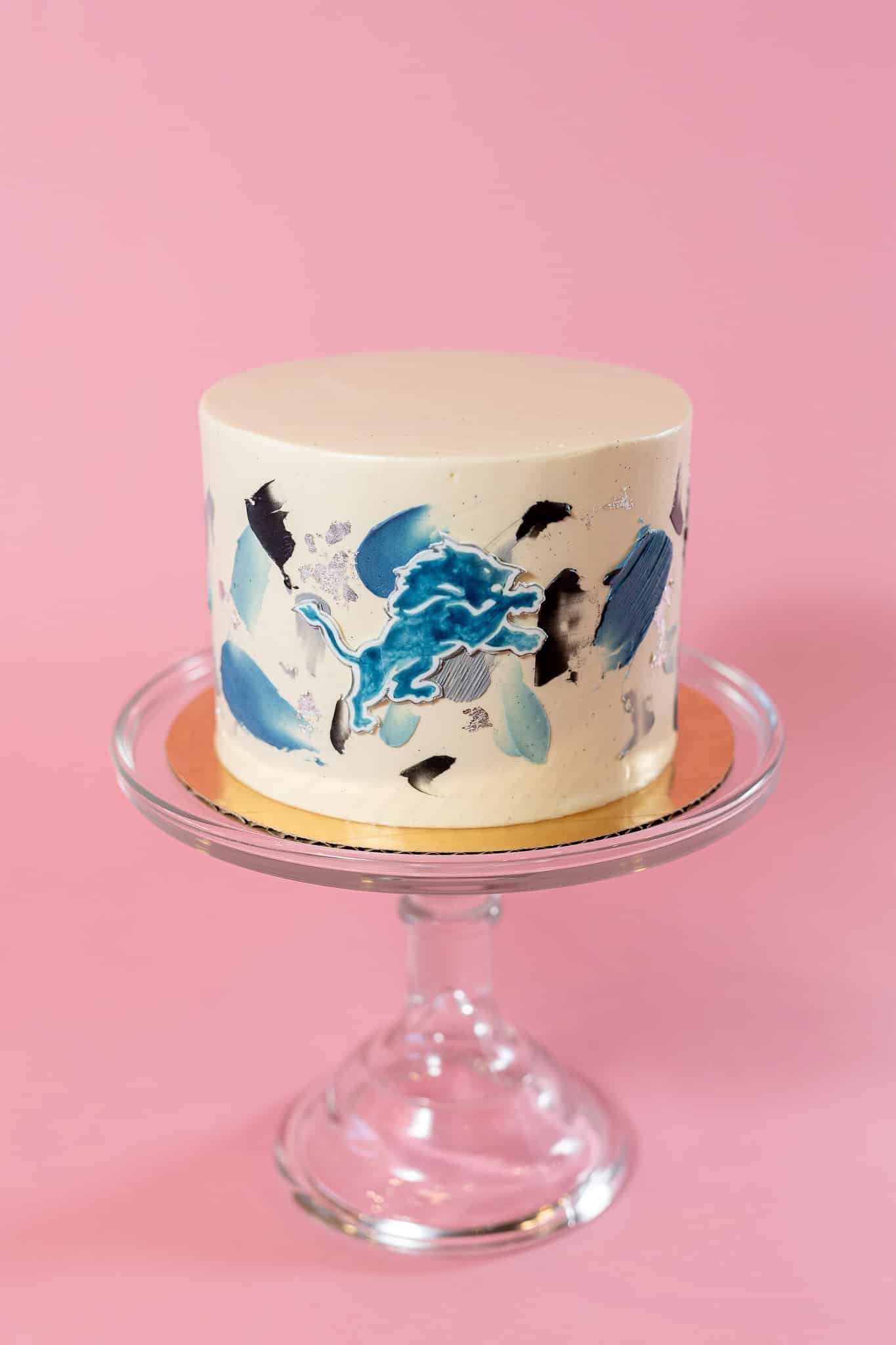 Cake with blue abstract design on glass cake stand, pink background.