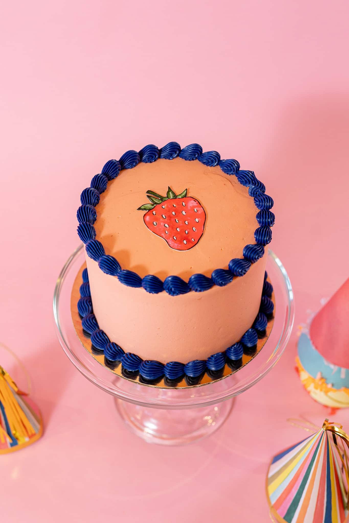 Strawberry cake with blue frosting detail, on a glass cake stand, pink background.