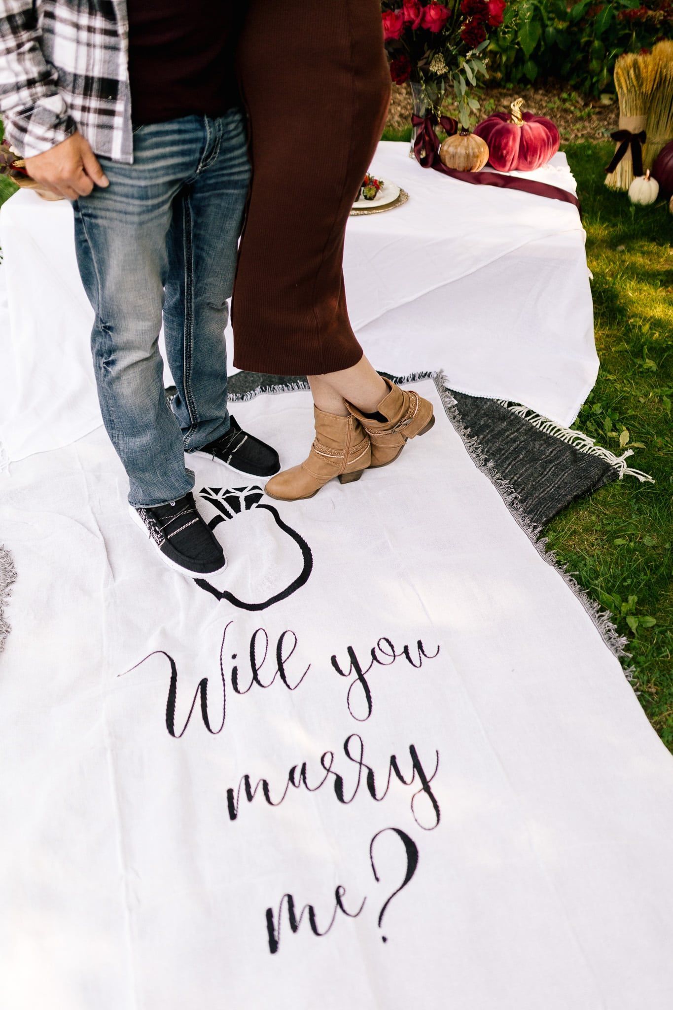 A man and woman are standing on a blanket that says `` will you marry me ''.