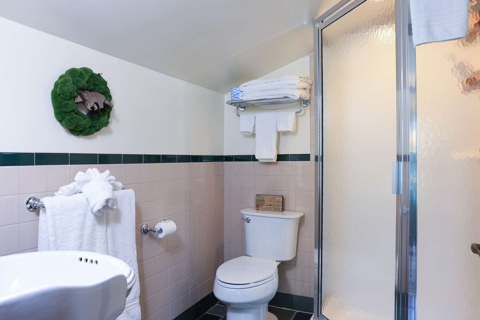 A bathroom with a toilet , sink , shower and wreath on the wall.