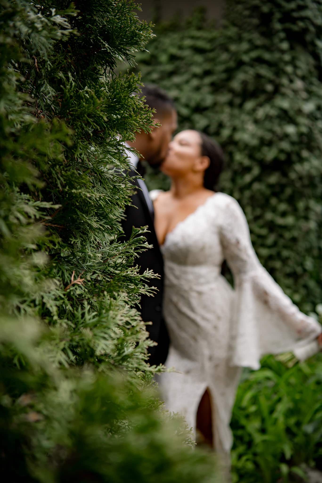 A bride and groom are kissing behind a bush . the bride is wearing a white dress.