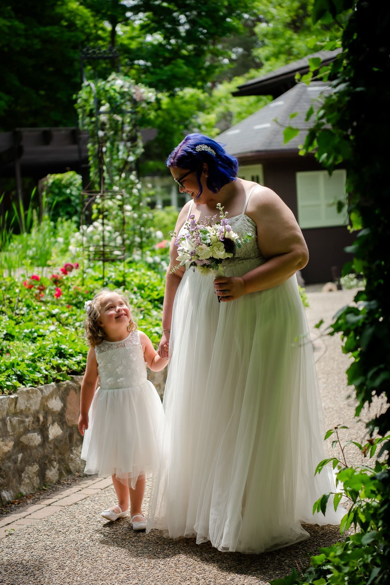 A bride and her flower girl are holding hands and walking down a path.