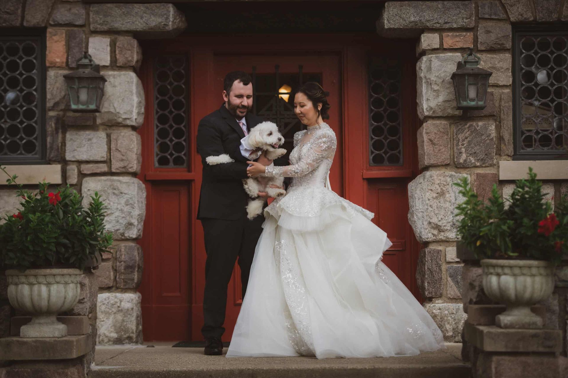 A bride and groom are holding a small white dog in front of a building.