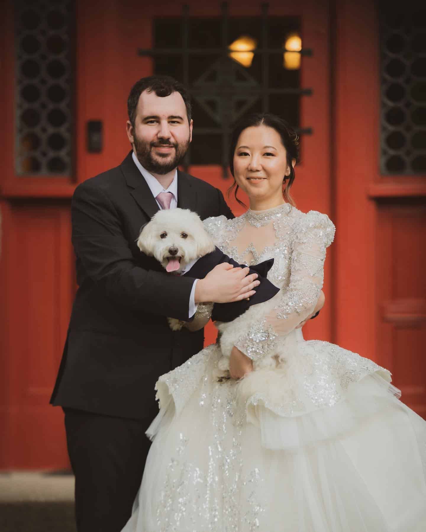 A bride and groom are holding a small white dog in front of a red door.