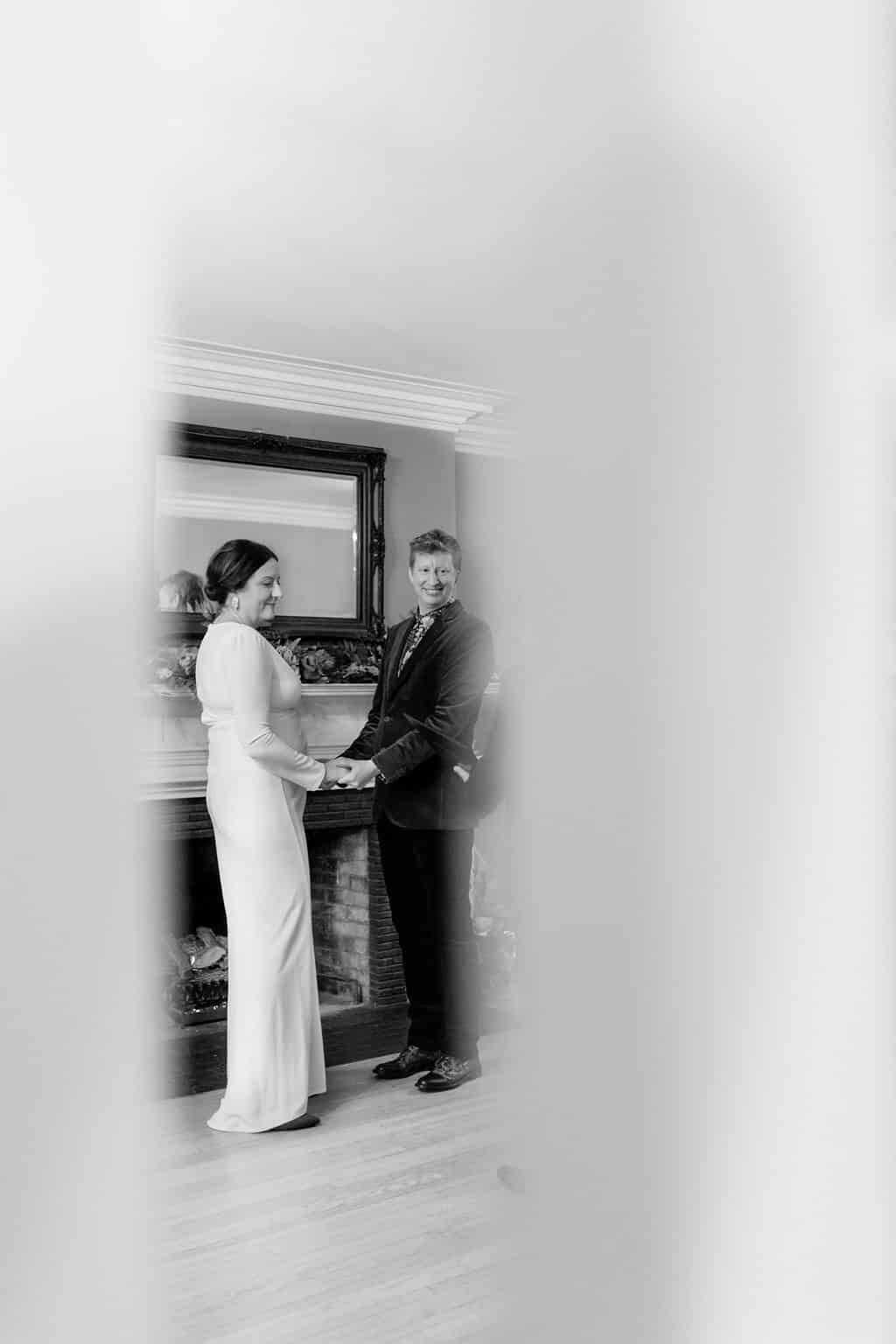A black and white photo of a bride and groom holding hands in front of a fireplace.