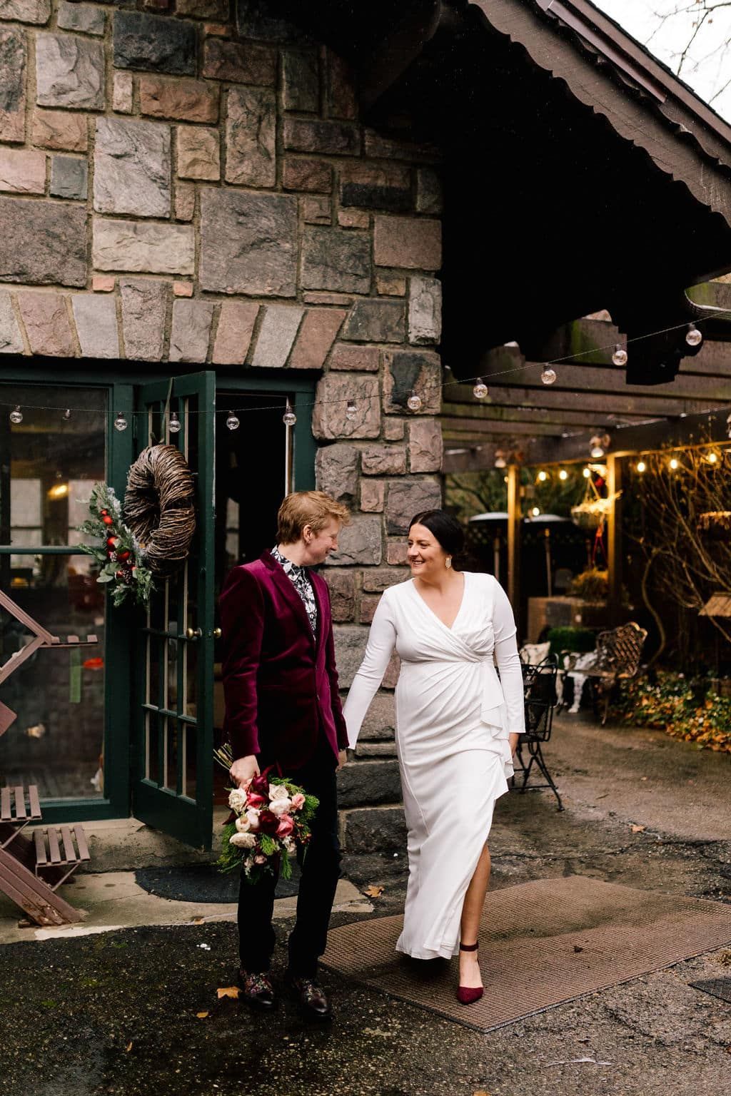 A bride and groom are walking in front of a stone building holding hands.