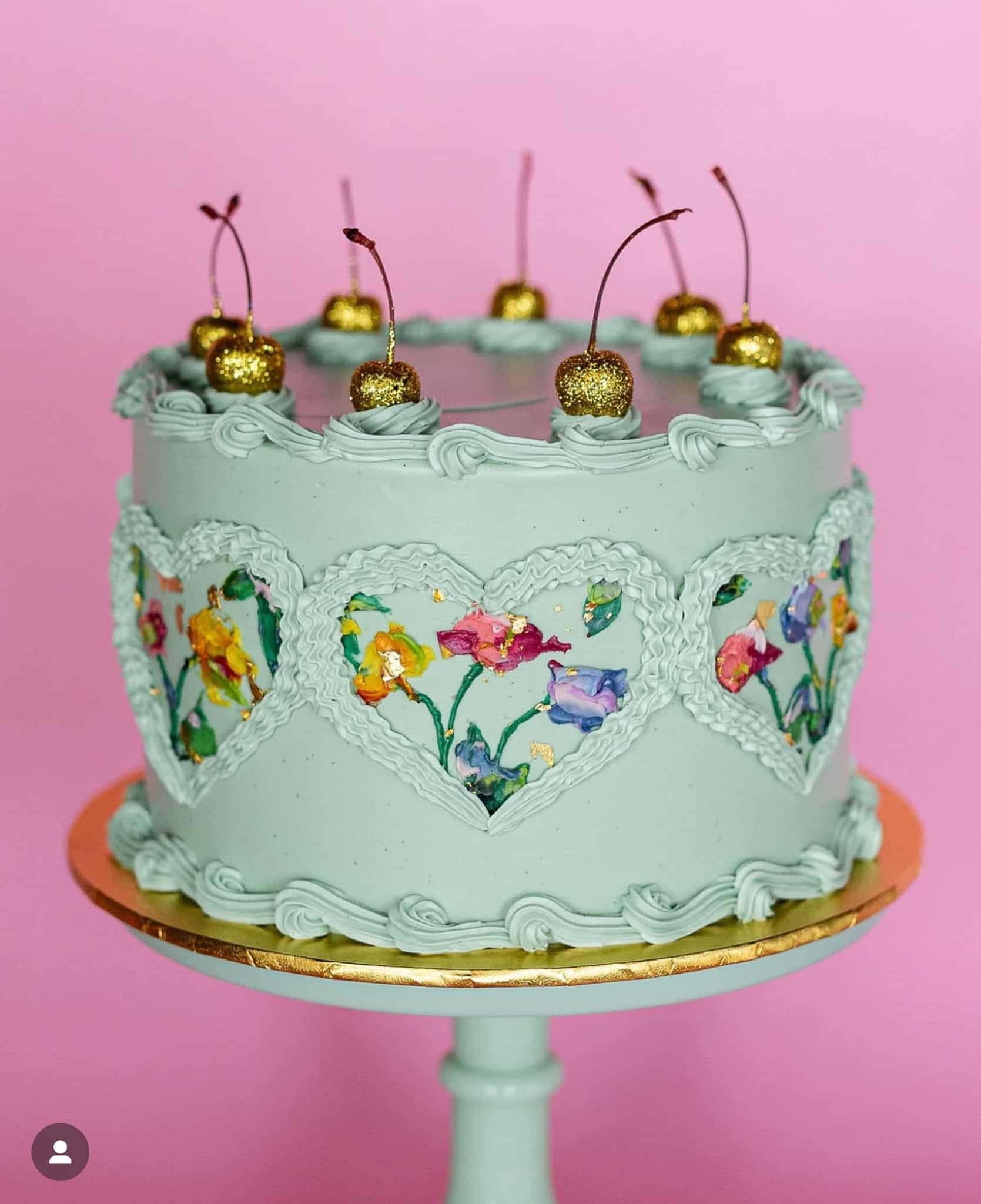 Mint green cake with floral heart designs, topped with gold-covered cherries, on a pink background.