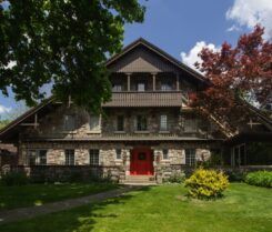 Large stone house with red door, balcony, and gabled roof, set in a green yard with trees under a blue sky.