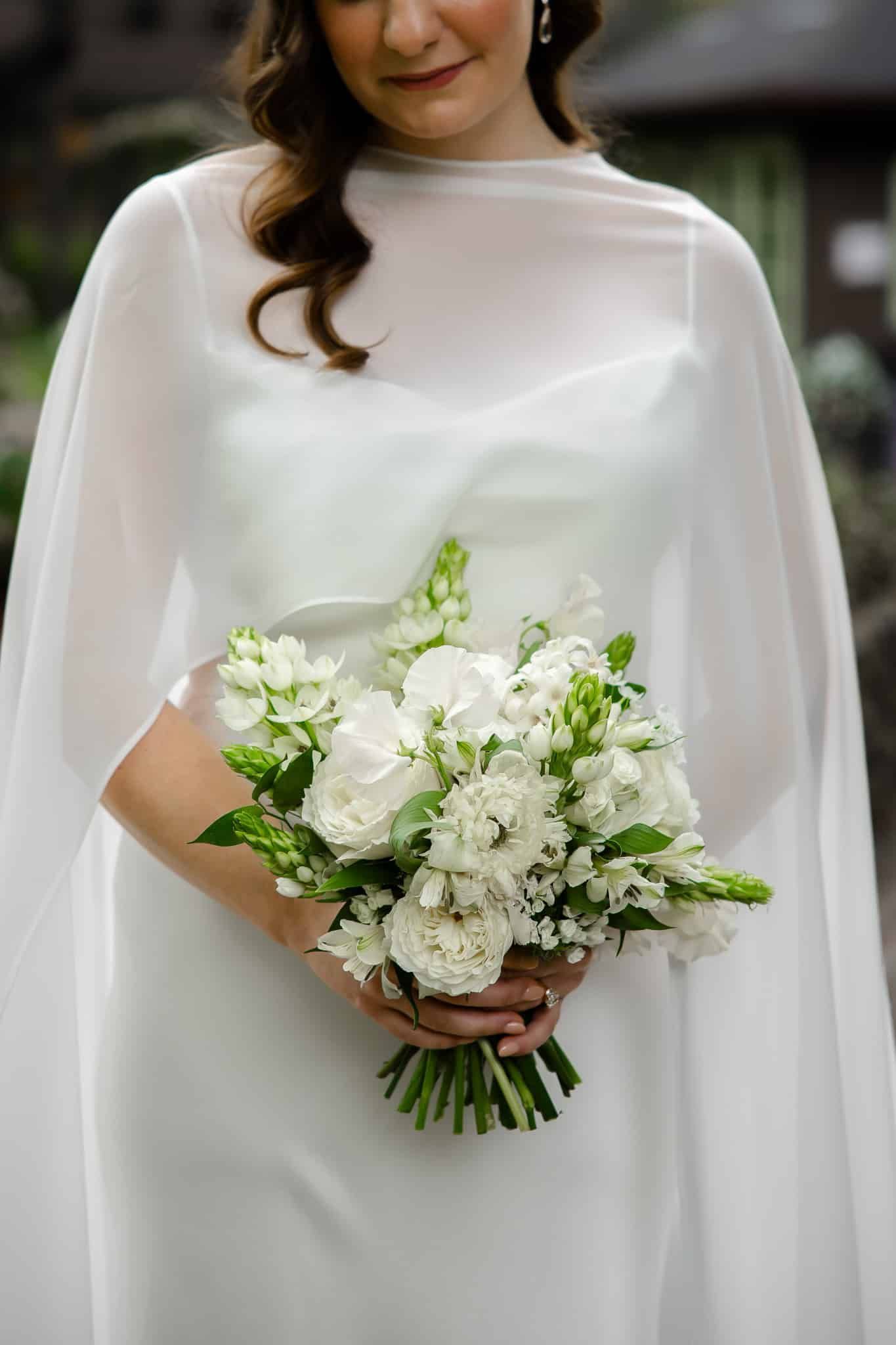The bride is wearing a white cape and holding a bouquet of white flowers.