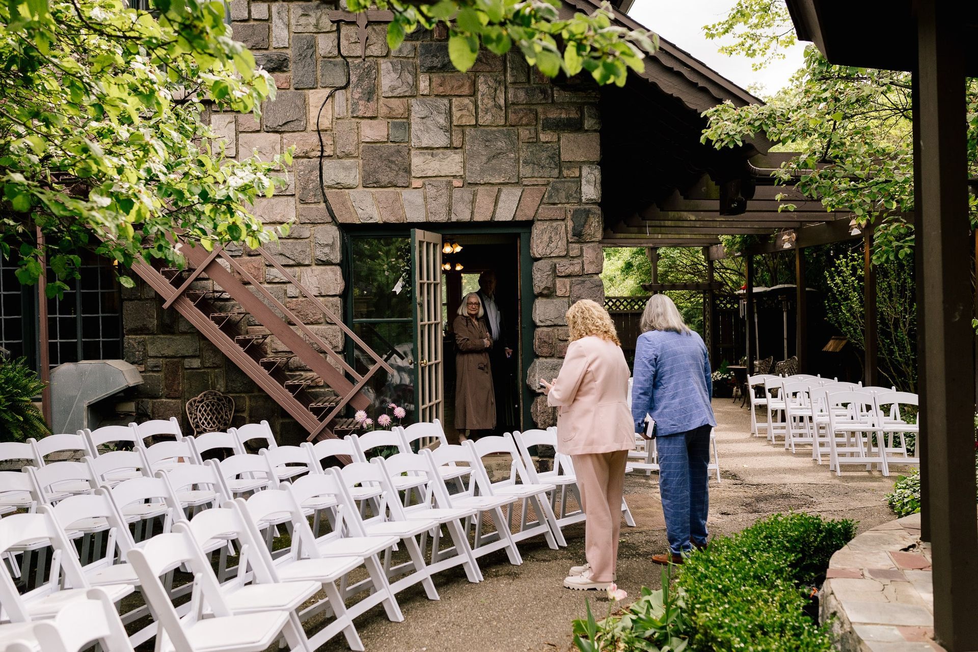 A couple of people standing in front of a stone building with rows of white chairs.