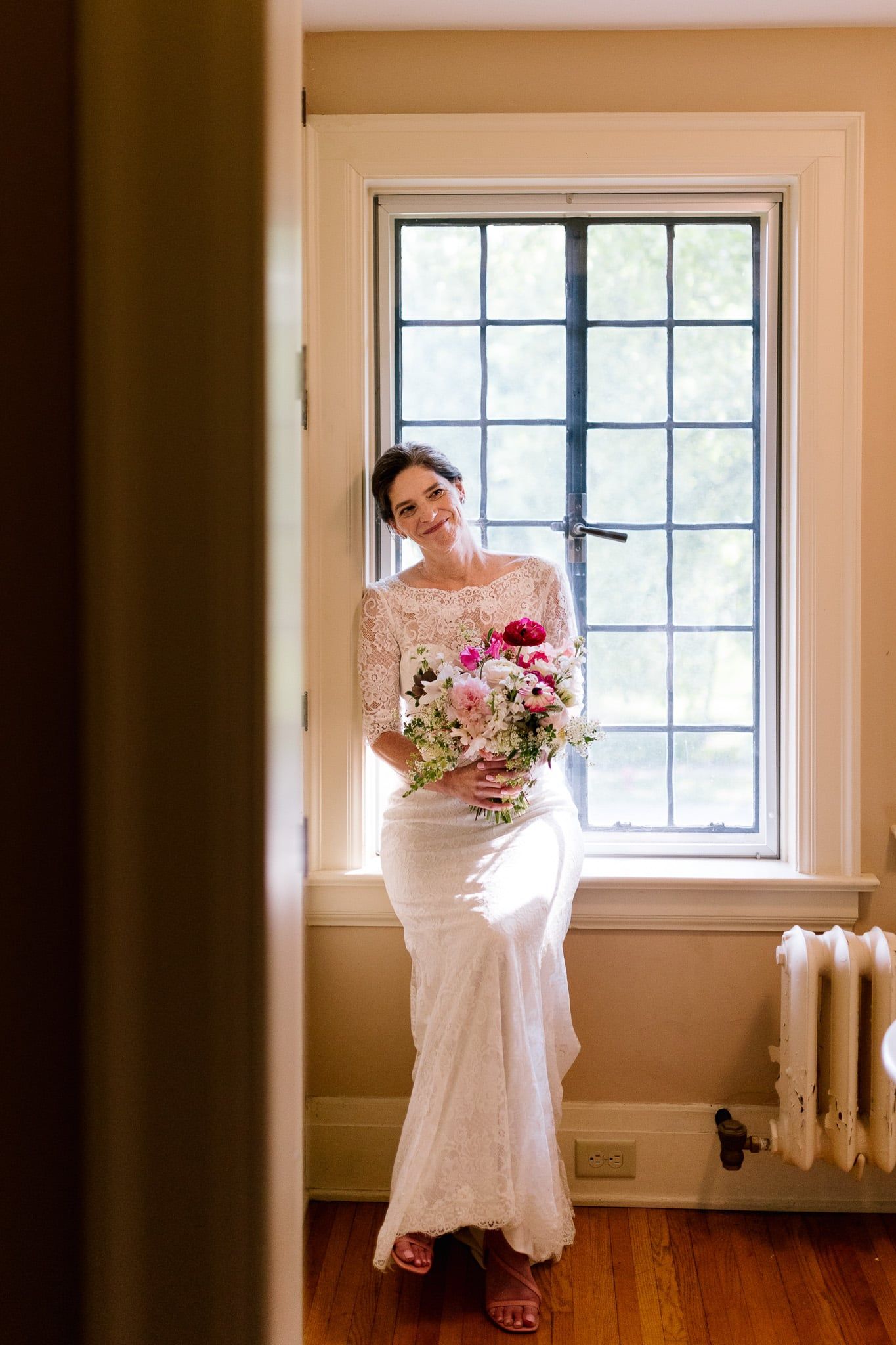 A woman in a wedding dress is standing in front of a window holding a bouquet of flowers.