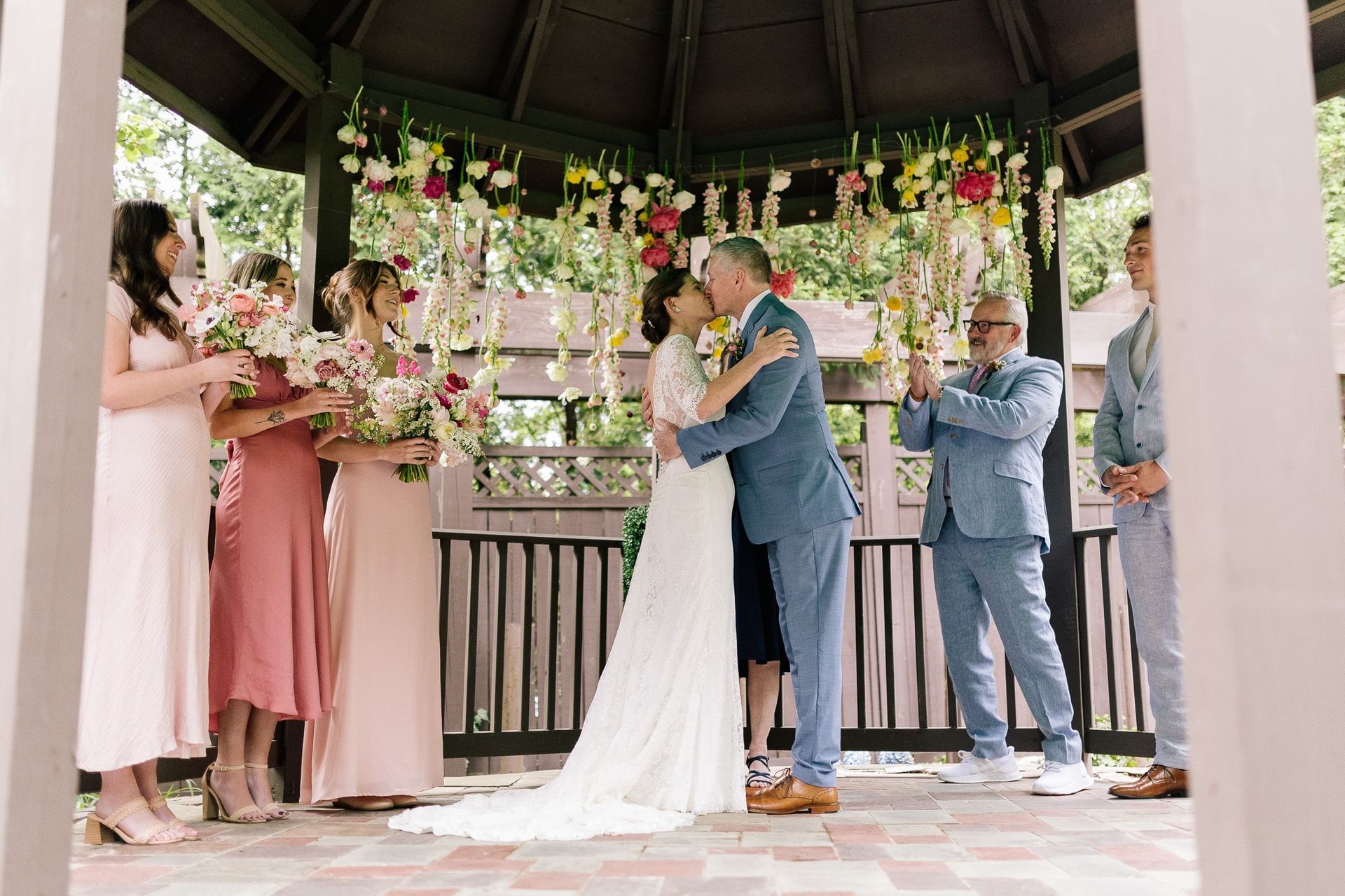 A bride and groom kissing in front of their wedding party under a gazebo.