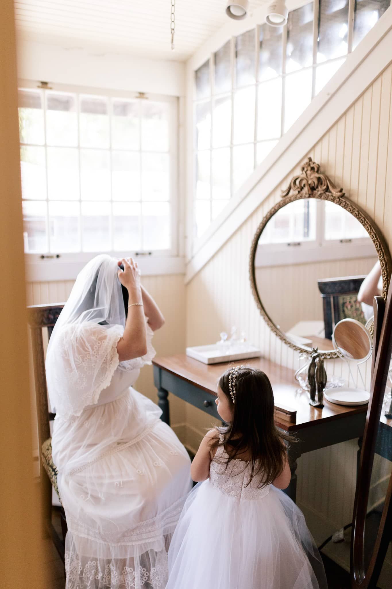A bride and a flower girl are getting ready for a wedding in front of a mirror.