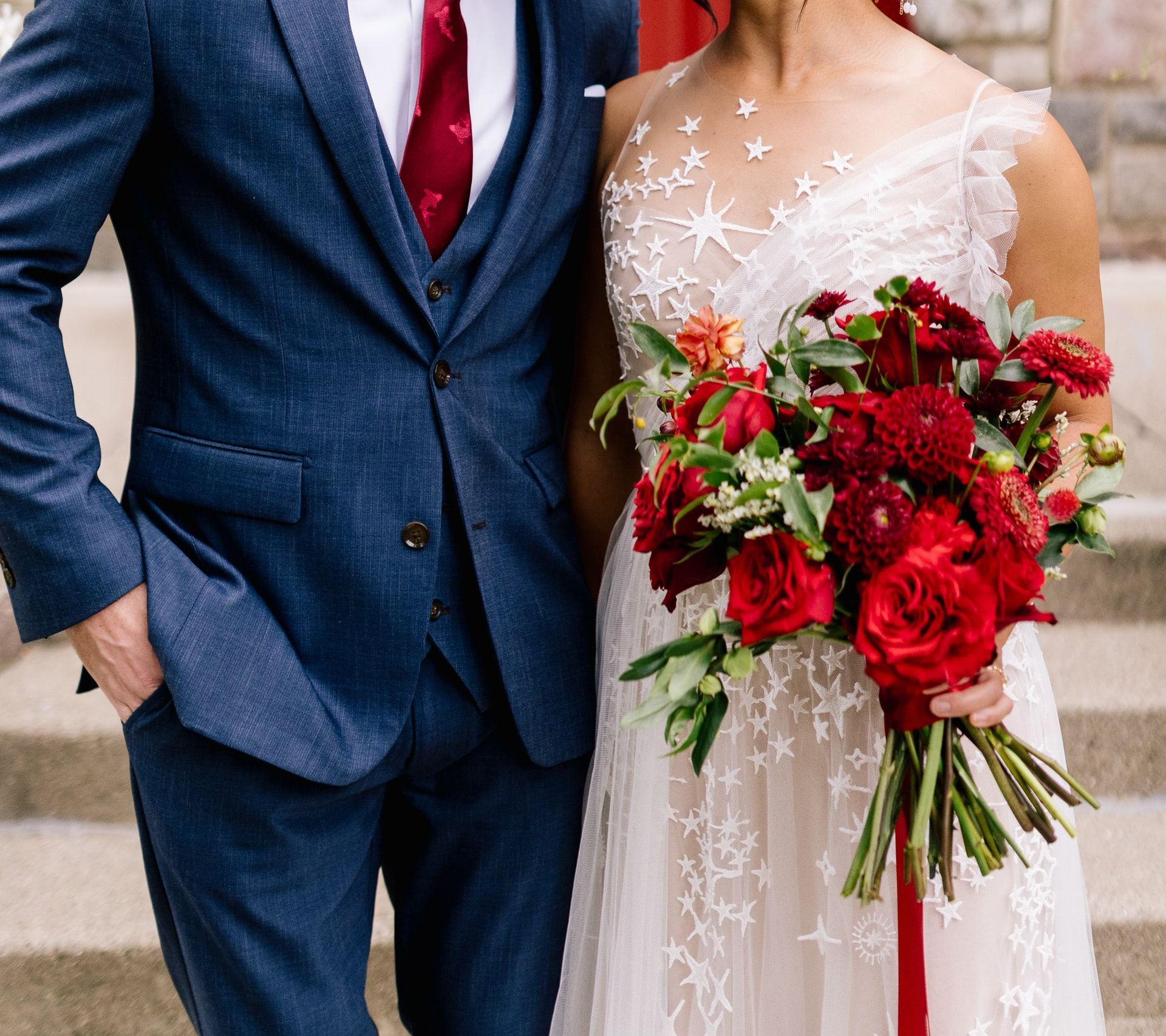 A bride and groom are posing for a picture while the bride is holding a bouquet of red flowers.