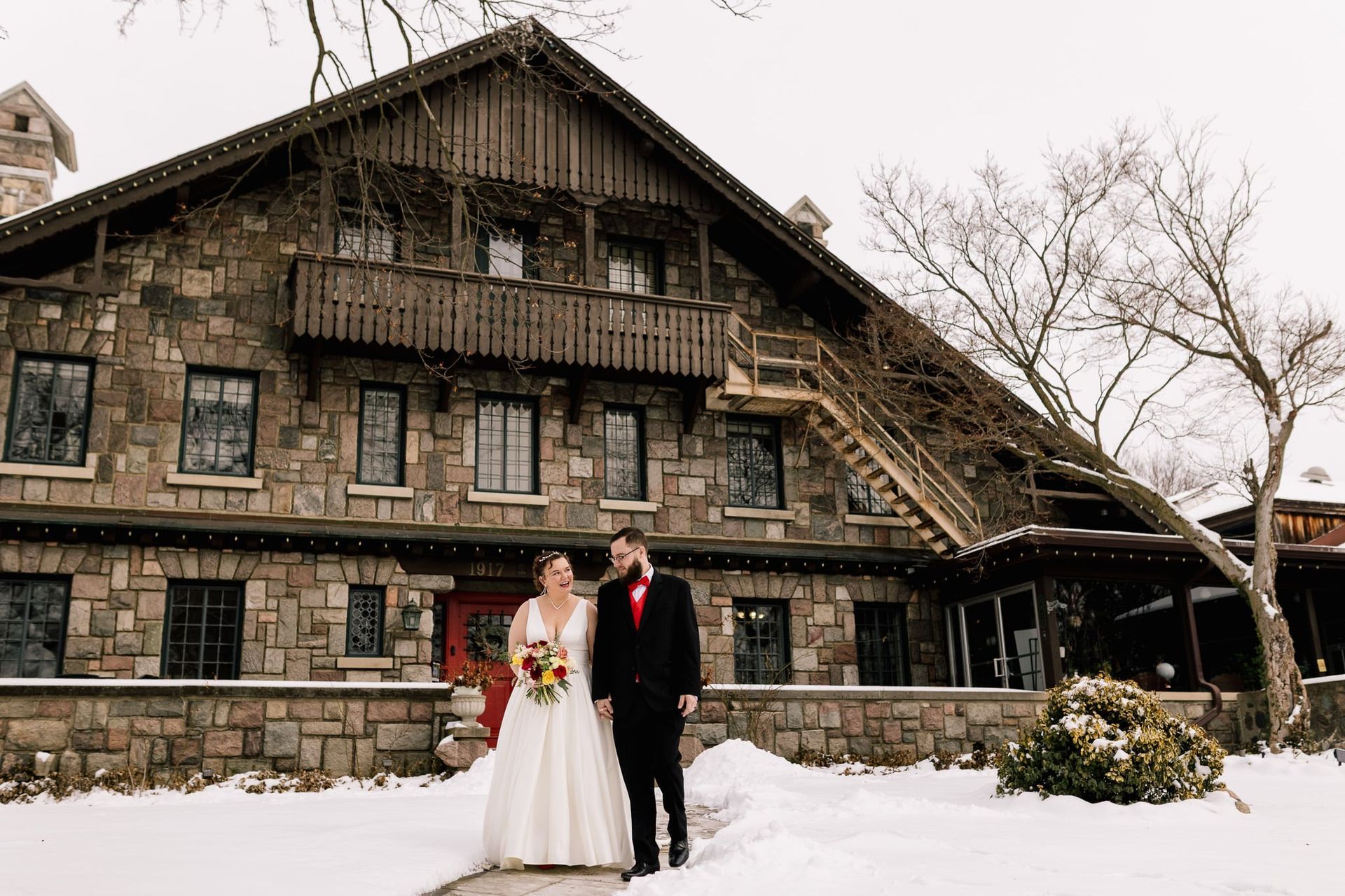 Bride and groom pose outside a stone lodge on a snowy day, holding hands.