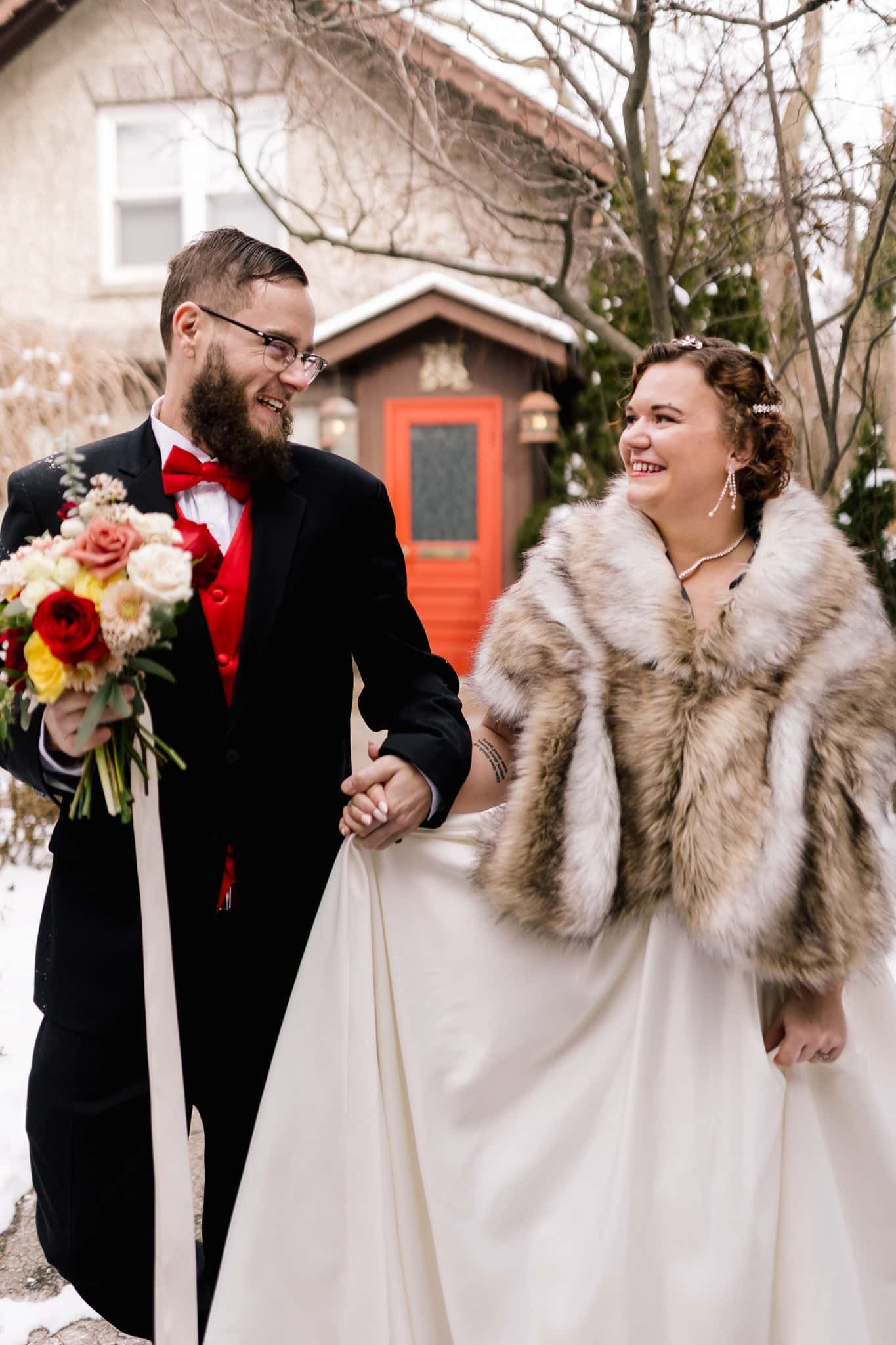 Bride and groom hold hands, walking. Bride wears fur shawl, groom holds bouquet. Red door and snow in background.